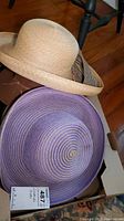 Two straw hats placed in a box, one light natural straw color, the other purple with spiral detailing.