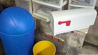 Group photo showing white mailbox, blue trash can with dome lid, and small yellow bin together in basement setting