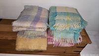 Seven folded mohair throw blankets on wooden surface showing pastel plaid patterns and fringed edges