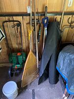 Photo of seed spreader, rakes, shovel, pitchfork, black rolled garden cloth, and white bucket leaning against barn wall