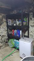 Full view of a black plastic shelving unit with various household items stored on it in a basement setting. Stone wall background visible.