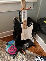 Black Fender Squier Bronco bass guitar with maple neck and fretboard, white pickguard, sitting on guitar stand. Guitar stand and guitar strings visible nearby.