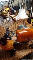Photo showing various lamps on a table including wall-mounted lamp, flexible desk lamp with metal base and white shade, heavy metal rooster lamp, and an orange-brown lamp body.