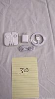 Two sets of Apple wired ear buds in their plastic cases, two Apple charging cubes, and two coiled Apple Lightning charging cables laid out on light fabric background.