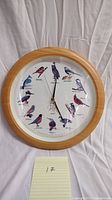 Photo of a round wall clock with bird illustrations and light wood-tone frame, showing all 12 birds and clock hands at center.