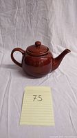Front view of brown ceramic teapot showing handle, body, lid, and spout on a plain fabric background with item number note.