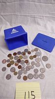 Coins spread on a fabric surface beside the blue plastic coin bank with its lid removed and placed nearby.