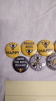 Photo of six bee-themed pins arranged in two rows on a cloth background. Pins have slogans 'HAPPY,' 'PRAISE BEES,' 'SAVE THE BEES PLEASE!,' 'MIND YOUR BEESWAX,' and images of bees.