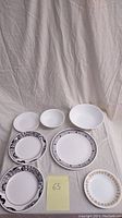 Seven Corelle dishware pieces arranged on cloth background, showing variety of plates and bowls with decorative borders