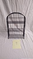 Front view of a small black metal rack with wire shelves and decorative scrollwork, placed against fabric backdrop with note '9' in front