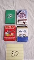 Four vintage playing card decks displayed face up showing front packaging: Saskatchewan Roughriders green deck, Bubba Gump Movie Trivia deck, Cards Eh! Canadian facts deck, and Pacific Pilsner deck.