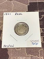 Front side of 1821 Mexico Spanish Colony silver real coin showing a crowned profile and inscriptions, in a white paper holder with handwritten notes.