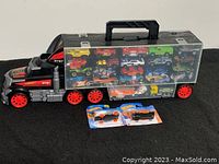 Overall view of the black and red plastic toy truck carrying case filled with over 50 colorful die-cast cars visible through clear panels; two packaged Hot Wheels cars in front.