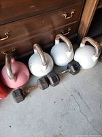 Photo of three kettlebells and two hex dumbbells on the floor in front of a wooden dresser; handles show rust and wear