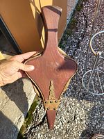 Photo of handmade wooden fire blower with metal rivets and brass decorative element, held by a hand outdoors on gravel ground.