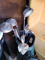 Golf club set in a green golf bag placed on a wooden floor against a wooden wall. Multiple clubs including irons and woods are visible.