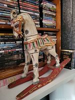 Vintage wooden rocking horse figurine with painted white body and red and gold accents placed on shelf with DVDs in background.