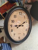 Photo of a large oval wall clock leaning against chair leg. Clock has a black textured frame, beige face with black numbers and hands, analog quartz movement.