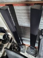 Photo of two tall floor-standing black speakers with fabric grille, standing on a dusty floor inside a storage space.