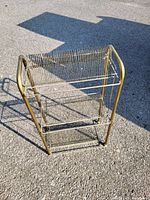 Photo of a gold-colored metal utility cart with two wire mesh shelves on an asphalt surface