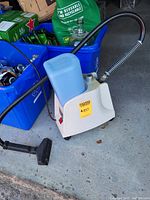 Portable carpet and upholstery cleaner with attached hose and cleaning head sitting on concrete next to recycling bins.