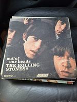 Stack of two Rolling Stones vinyl records on a car seat, top showing 'Out of Our Heads' album cover with band members' faces and London Records logo.