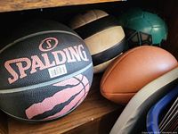 Photo showing four sports balls on a shelf: Spalding basketball, black and white volleyball, brown football, green and black soccer ball