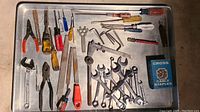 Various hand tools including wrenches, screwdrivers, pliers, files, tape measure, and a box of cable staples arranged on a metal tray.