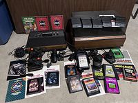 Full display of Atari CX-2600A console and all included accessories spread around it on a dark surface. Includes 2 joysticks, 2 paddles, push button, power cord, TV switch, 14 game cartridges, original game center storage case and catalogs.