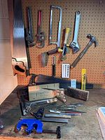 Full view of the hand tools displayed on pegboard and workbench including pipe wrenches, saws, mitre box, hammer, wire brush, clamps, and assorted small tools.