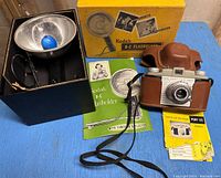 Photo of Kodak B-C Flashholder with reflector, flash mounting component inside original blue box, original yellow Kodak B-C Flashholder box, Kodak Pony 35mm camera in brown leather case with strap, and instruction manuals for both flashholder and camera.