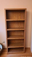 Front view of tall wooden bookcase with five shelves showing oak laminate finish and backing.