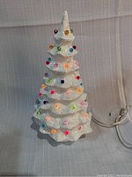 White ceramic Christmas tree decorated with small, colorful bulbs that light up. Tree has several tiers with bulb placements spread evenly across the surface. The electrical cord and plug are visible, indicating the tree lights up.