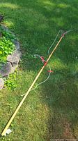 Photo showing the long wooden pole tree trimmer with pruner and pull cords lying on grass near garden rocks.