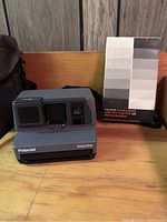 Polaroid Impulse instant camera and unopened black and white Type 105 film pack sitting on wooden surface with wooden backdrop, showing camera front with flash unit and film pack box's grayscale gradient design.