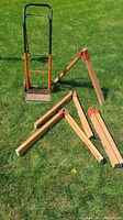 Photo showing four wooden saw horses partially assembled with red metal clamps and one metal appliance dolly with black handles and orange frame on grass.