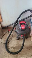 Photo of small black and red Shop Vac vacuum with hose and wand on a concrete floor.