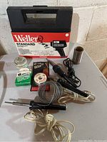 Overall photo showing Weller Standard soldering gun kit box, two soldering irons, solder wire, solder paste, and spool of wire.