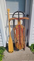 Photo showing a vintage wooden rowing oar labeled 'DAN' alongside a wooden sled with metal runners labeled 'Electric Navigator' and a vintage Little League baseball bat.