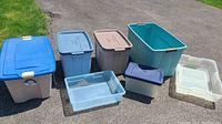 Lot of eight assorted plastic storage bins of varying sizes and colors displayed outdoors on pavement with grass nearby.