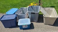 Seven assorted plastic storage totes and bins on pavement with grass background, showing different sizes and lid presence.