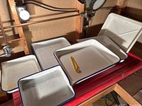 Photo showing multiple white enamel trays with blue rims and yellow plastic tongs inside, arranged on a red plastic tray in a darkroom setting.