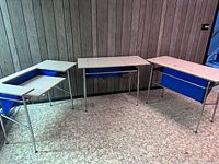 Three vintage audiovisual school desks arranged along wall, showing white tops, blue adjustable sections, and blue fabric storage pouches.