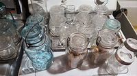 Over 25 vintage and antique canning jars arranged on a surface. Includes clear and blue glass jars with wire clamp lids and metal lids.