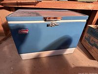 Exterior view of vintage Coleman metal cooler with dark jade turquoise exterior and white bottom, featuring a lockable latch and Coleman logo badge.