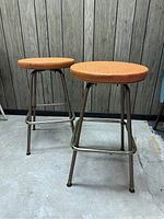 Two vintage MCM stools with round orange vinyl seats and metal frames, standing on concrete floor against wood panel wall.