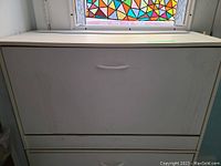 White shoe rack cabinet with two compartments shown from top angle with window and stained glass film in background