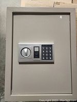 Front view of the closed grey steel in wall safe showing electronic keypad and turn handle.