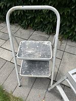 Metal two-step stool with worn paint and textured anti-slip steps, standing on stone pavement with green foliage in the background.