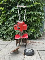 Front view of red Mantis roto tiller showing 8 inch tines, metal edger attachment, and detachable wheel on paved surface with green ivy background.